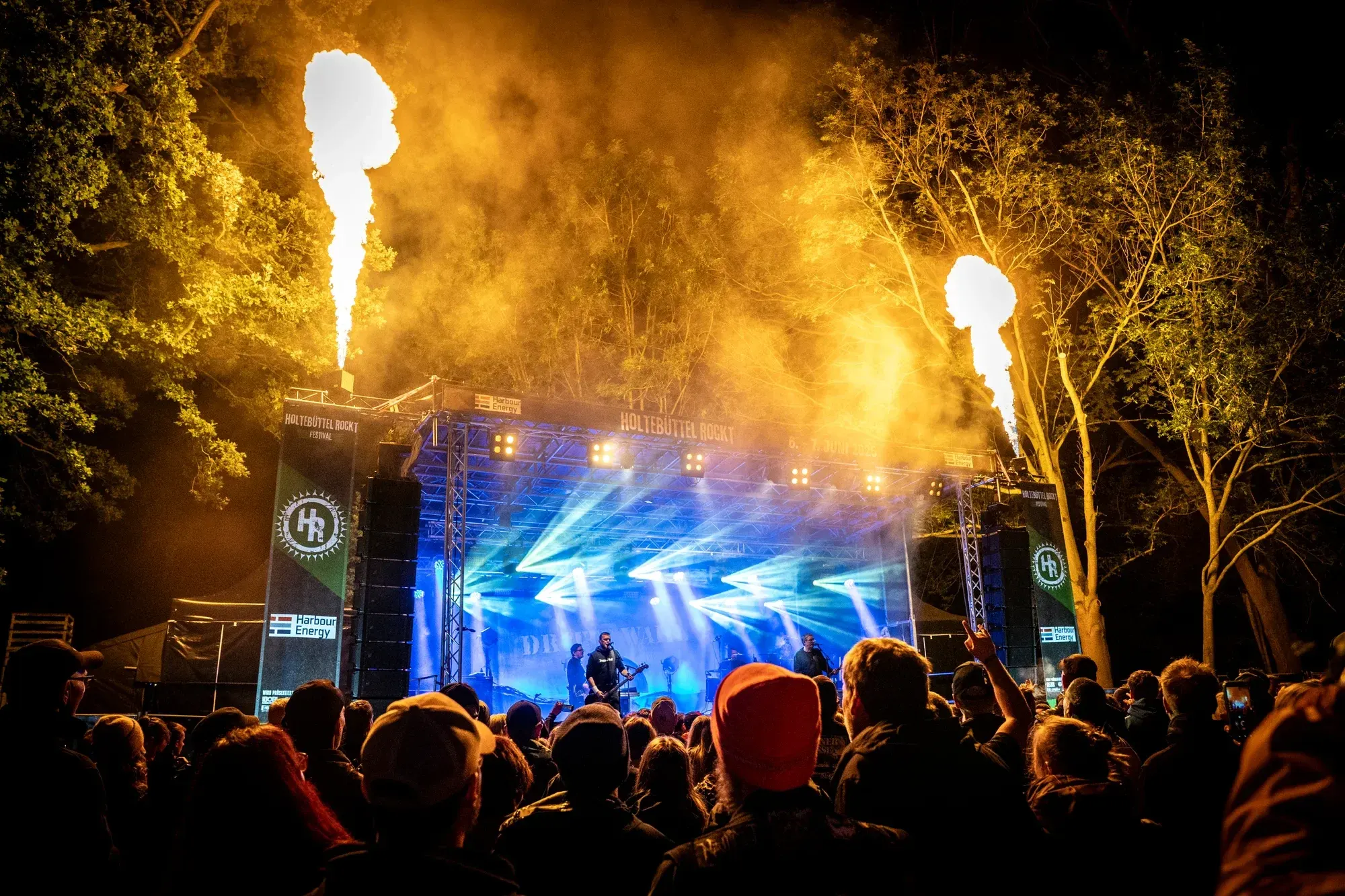 Publikum vor der blau beleuchteten Bühne mit Flammenfontänen beim Holtebüttel Rockt 2025
